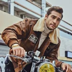 Homme sur une moto classique, affichant un blouson en cuir marron avec des patchs, look décontracté et robuste.