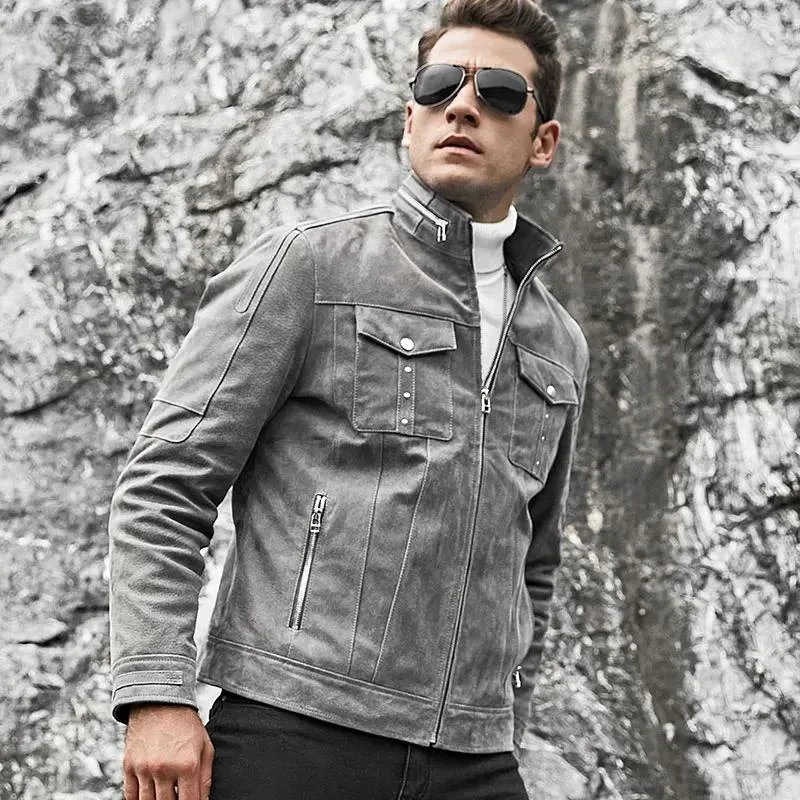 Homme stylé avec un blouson en cuir gris aspect vieilli et des lunettes de soleil, posant devant un mur de pierres. Idéal pour un look biker vintage.