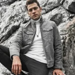 Homme assis dans un paysage rocheux, affichant son blouson en cuir gris avec un collier. Une veste de moto vintage pour un équipement motard complet.